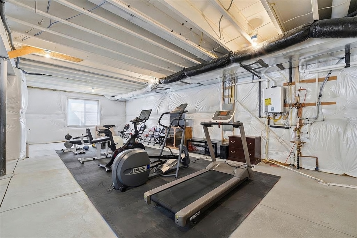 This unfinished basement space has been converted into a functional home gym, featuring a variety of exercise equipment including a Precor treadmill and elliptical machine. The room has exposed ceiling joists, concrete flooring, and walls covered in protective plastic sheeting, suggesting a utilitarian or workshop-style aesthetic. The perspective is a wide-angle shot from the corner of the room, capturing the full layout of the workout area.