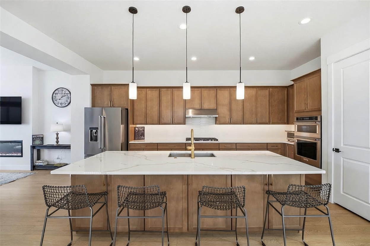 This modern, open-concept kitchen features warm wood cabinetry, a large central island with a white quartz countertop, and a gold-toned faucet. Three sleek pendant lights hang above the island, which is paired with four dark metal bar stools, creating a sophisticated and inviting space. The kitchen is fully equipped with stainless steel appliances, including a built-in oven and microwave, and offers a clean, contemporary aesthetic.