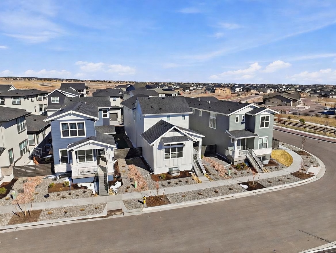 This high-angle aerial view captures a suburban neighborhood featuring a row of modern, multi-story single-family homes with varied siding colors and architectural styles. The scene highlights the clean, paved streets, landscaped front yards with gravel beds, and the surrounding residential development under a clear blue sky. The perspective provides a comprehensive look at the property's placement within the community and its proximity to neighboring houses.