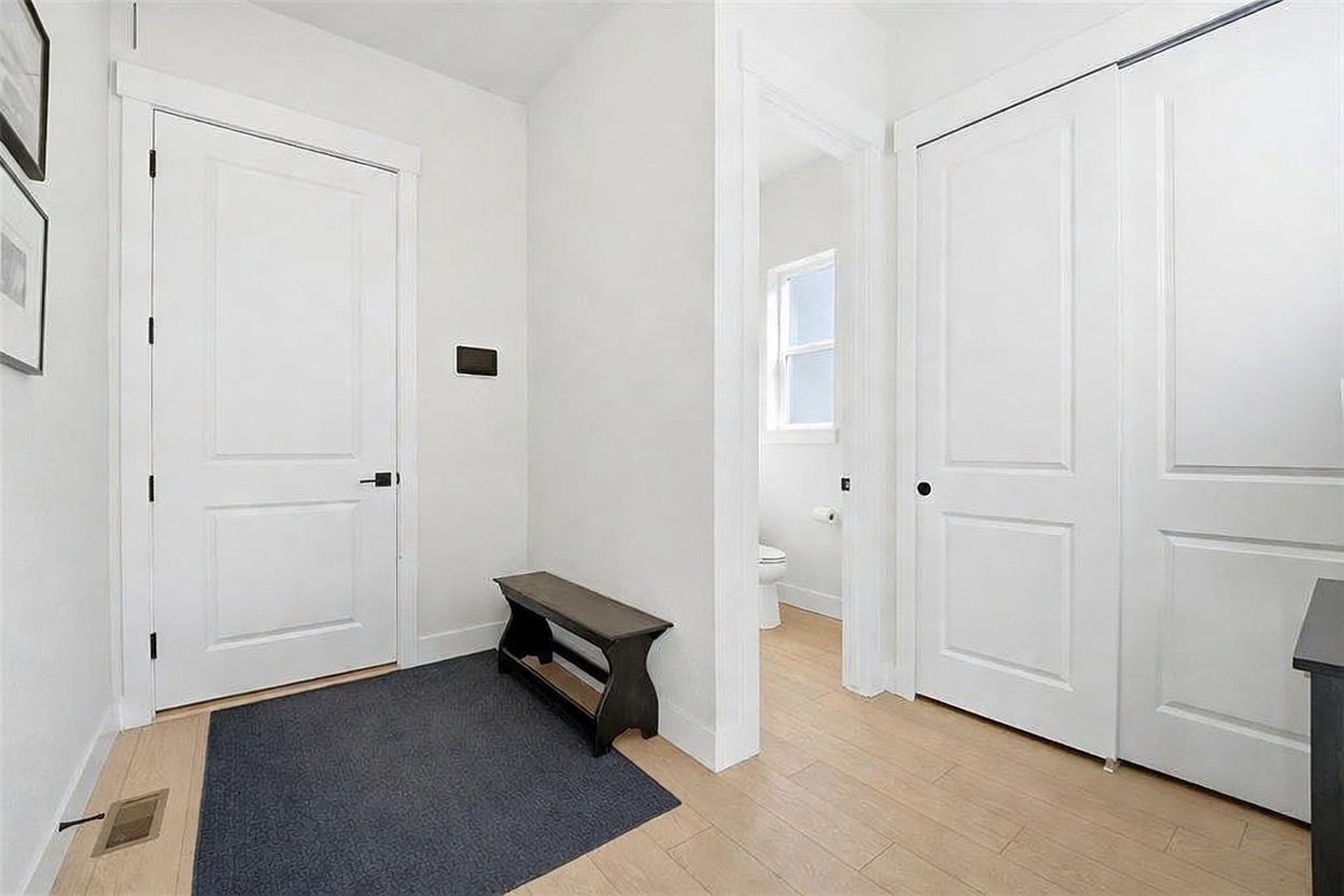 This bright and modern hallway features light wood-look flooring and crisp white walls, creating a clean and airy atmosphere. The space includes a dark wooden bench placed on a charcoal rug, a doorway leading to a bathroom, and a set of double doors, likely a closet. The perspective is eye-level, capturing the functional layout and minimalist aesthetic of the home's interior.