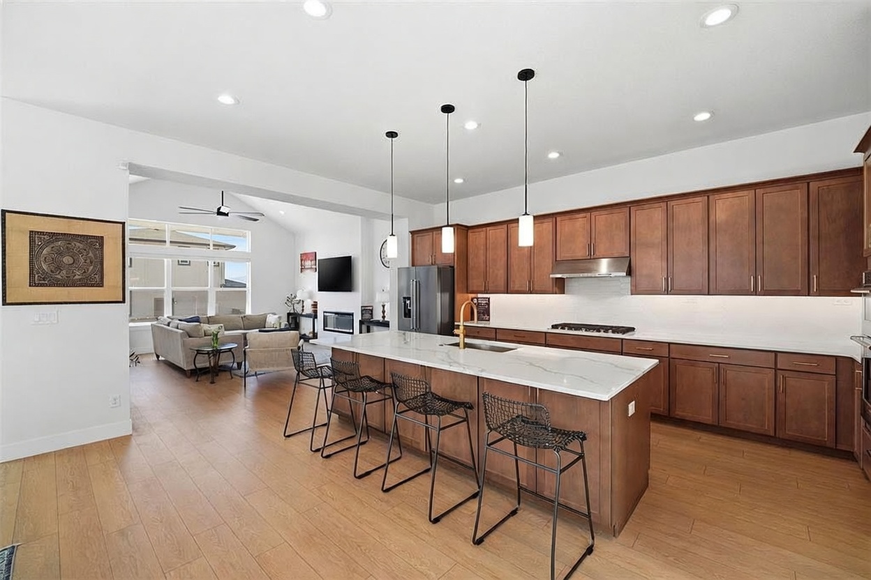 This spacious, open-concept kitchen features warm wood cabinetry, a large central island with a white quartz countertop, and modern black metal bar stools. The area is illuminated by three sleek pendant lights and recessed ceiling lighting, creating a bright and inviting atmosphere that flows seamlessly into the adjacent living room. The design is contemporary and functional, highlighting a clean aesthetic with high-end finishes.