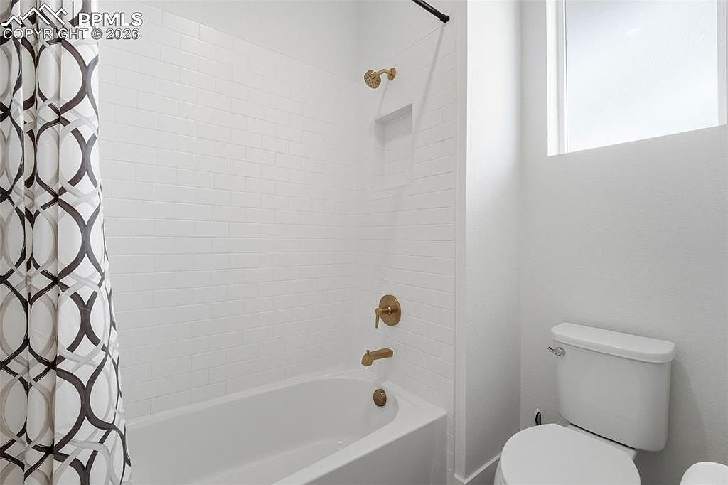 This clean and modern bathroom features a white subway-tiled shower surround with a built-in niche and gold-toned fixtures. A white porcelain toilet sits adjacent to the tub, while a patterned shower curtain adds a touch of visual interest to the bright, neutral space. The perspective is a standard eye-level shot capturing the functional layout of the bathroom.