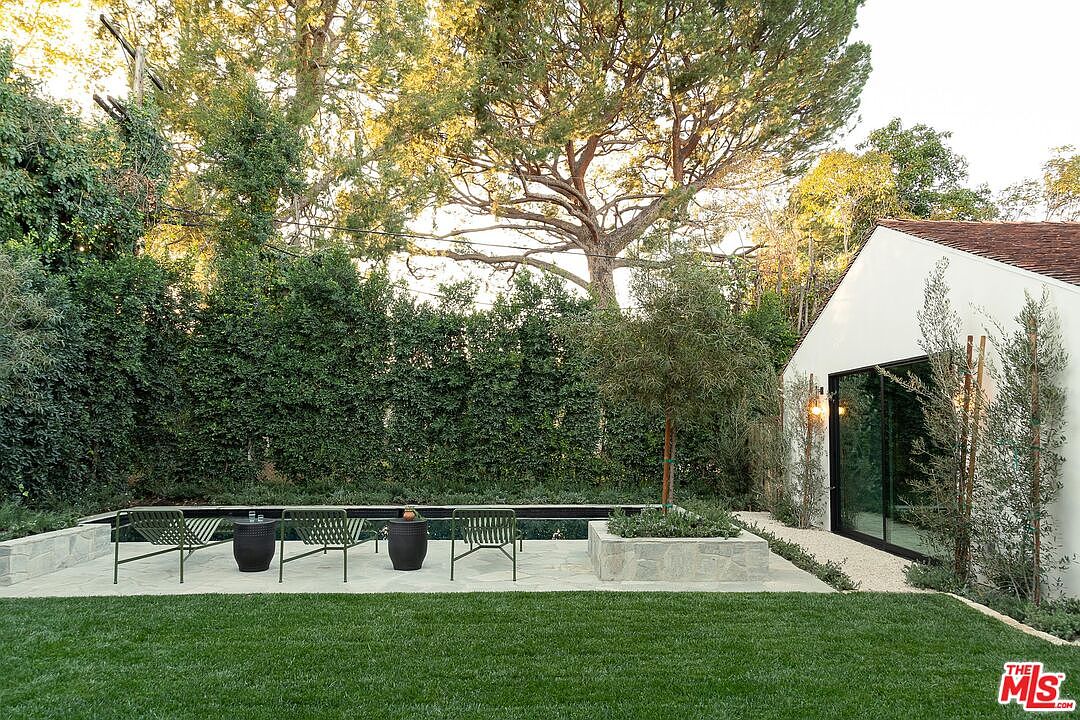 This exterior shot showcases a beautifully landscaped backyard featuring a sleek, dark-bottomed pool, modern outdoor furniture, and lush green lawn. A tall, manicured hedge provides privacy, while a contemporary white building with large glass doors adds architectural interest. The overall impression is one of tranquility and sophisticated outdoor living.