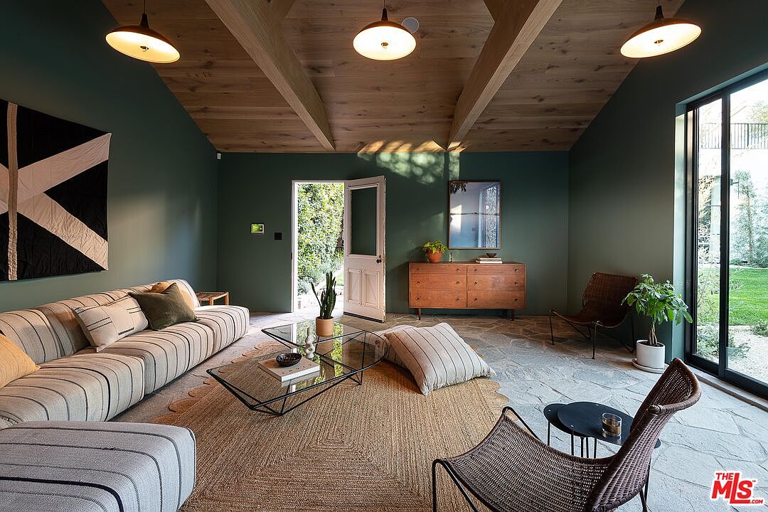 This is an interior shot of a living room featuring dark green walls, a wooden beamed ceiling, and stone flooring. The room is furnished with a striped sofa, a glass coffee table, and woven chairs, creating a cozy and stylish atmosphere. Natural light floods the space through a large glass door, offering a view of the outdoor greenery.