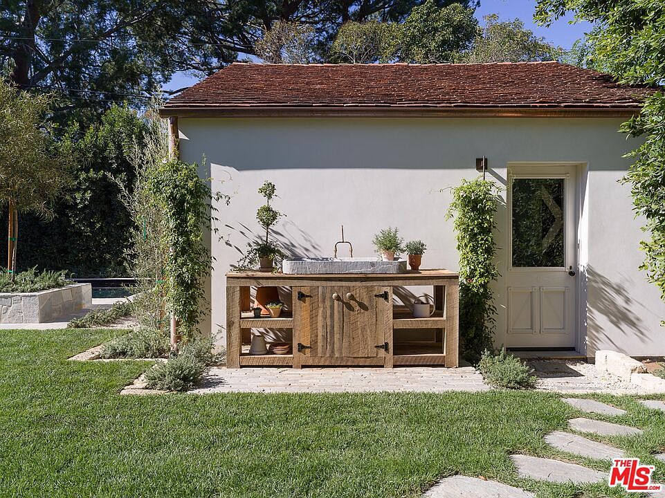 This image showcases a charming outdoor space featuring a rustic wooden sink station against a light-colored building with a red tile roof. The area is surrounded by lush greenery, including trees and climbing plants, with a well-maintained lawn and stone pathway adding to the inviting atmosphere. The scene evokes a sense of tranquility and is ideal for outdoor entertaining or relaxation.