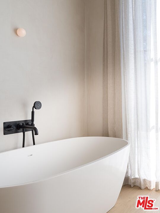 This image showcases a modern primary bathroom featuring a freestanding white bathtub with a sleek, matte black faucet and shower fixture. The walls are a neutral tone, complemented by sheer curtains that filter natural light. The overall aesthetic is minimalist and serene, creating a spa-like atmosphere.