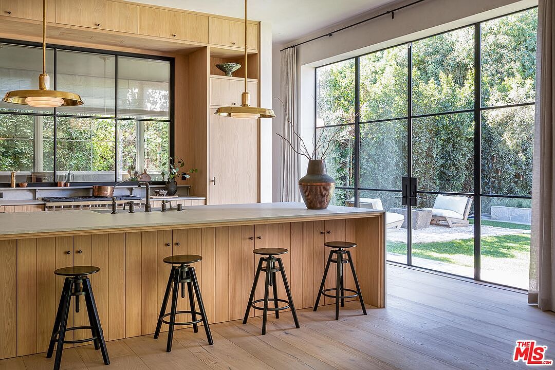 This is a bright and modern kitchen featuring light wood cabinetry and a large island with seating. The kitchen boasts a large window overlooking a lush garden, bringing natural light and a sense of tranquility to the space. Black framed windows and pendant lighting add a touch of contemporary elegance.