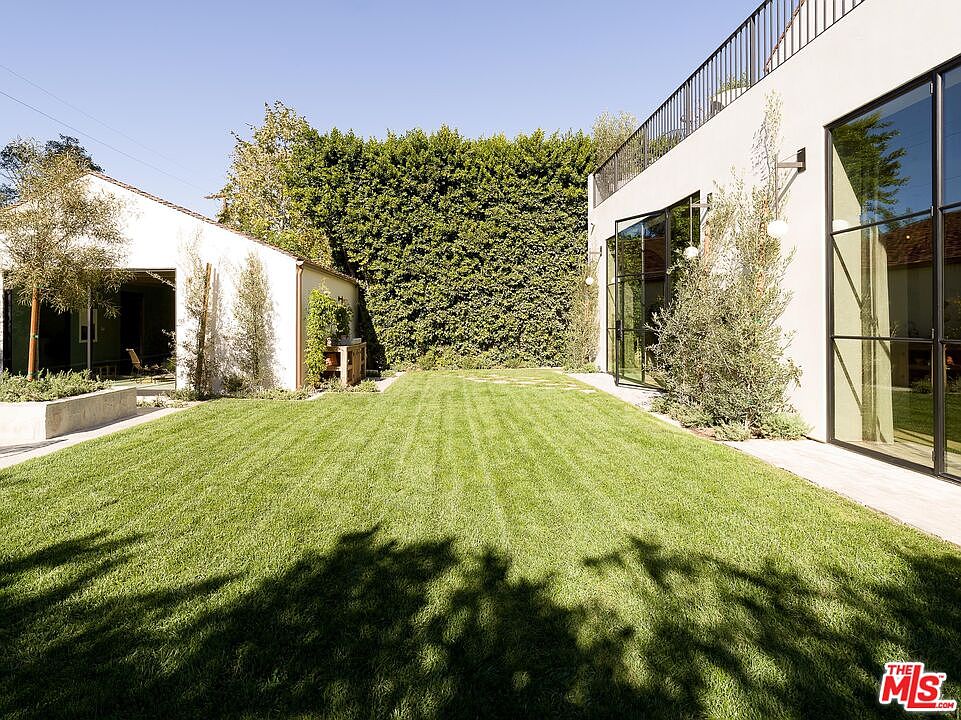 This image showcases a well-maintained backyard featuring a lush green lawn, a tall hedge providing privacy, and modern architectural elements. The yard is bordered by a building with large windows and a separate structure, creating a serene and inviting outdoor space. The perspective is a wide shot, capturing the breadth of the yard and its connection to the surrounding buildings.