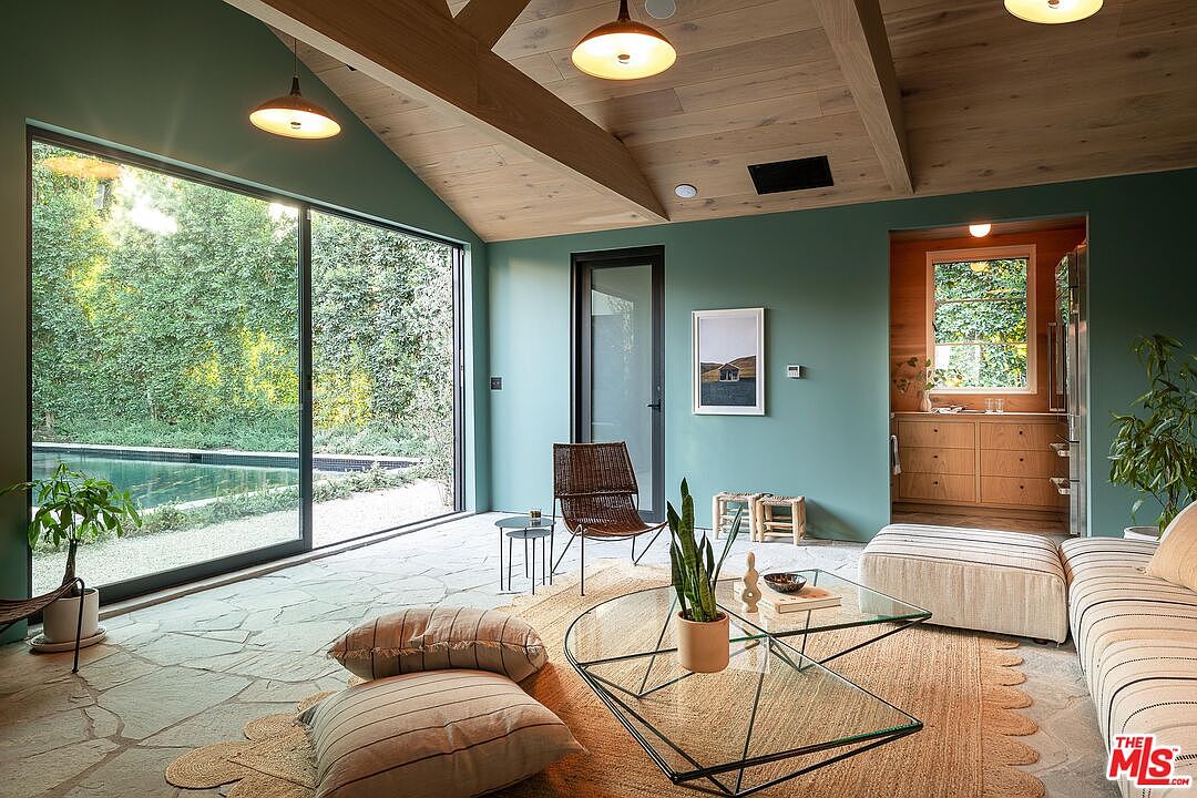 This interior shot showcases a modern living room with a seamless indoor-outdoor transition. The room features a large window overlooking a pool and lush greenery, complemented by a stone floor and a light wood ceiling with exposed beams. The decor includes a glass coffee table, woven rug, and neutral-toned furniture, creating a serene and inviting atmosphere.