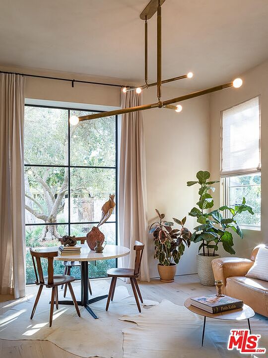 This interior shot showcases a stylish dining area with a round table set for two, complemented by a modern chandelier. Large windows provide ample natural light and a view of the outdoors, while potted plants add a touch of greenery. The room exudes a sophisticated and inviting atmosphere, perfect for intimate gatherings.
