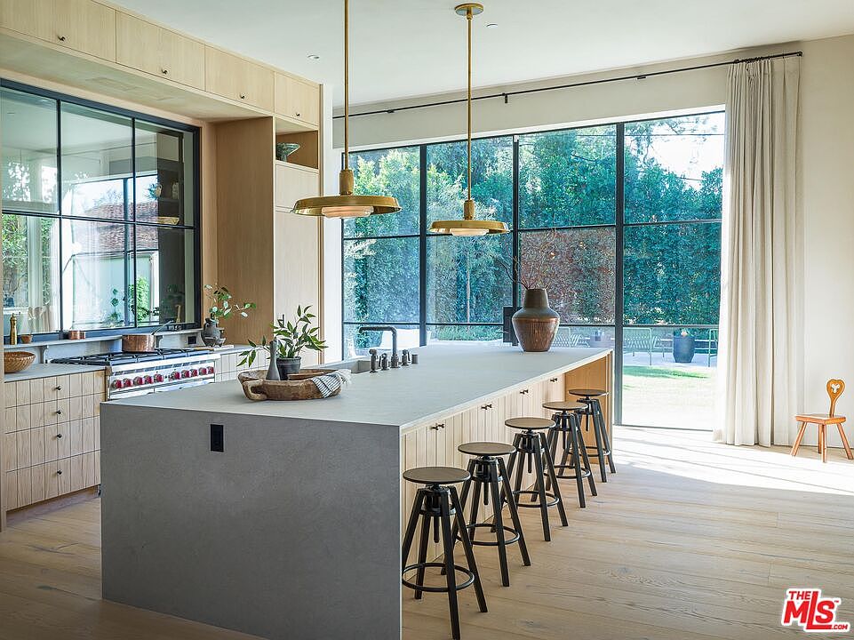 This is a bright and modern kitchen featuring light wood cabinetry and a large center island with seating. The kitchen is well-lit by natural light streaming through a large window overlooking the backyard. The design is clean and contemporary, emphasizing natural materials and a seamless connection to the outdoors.