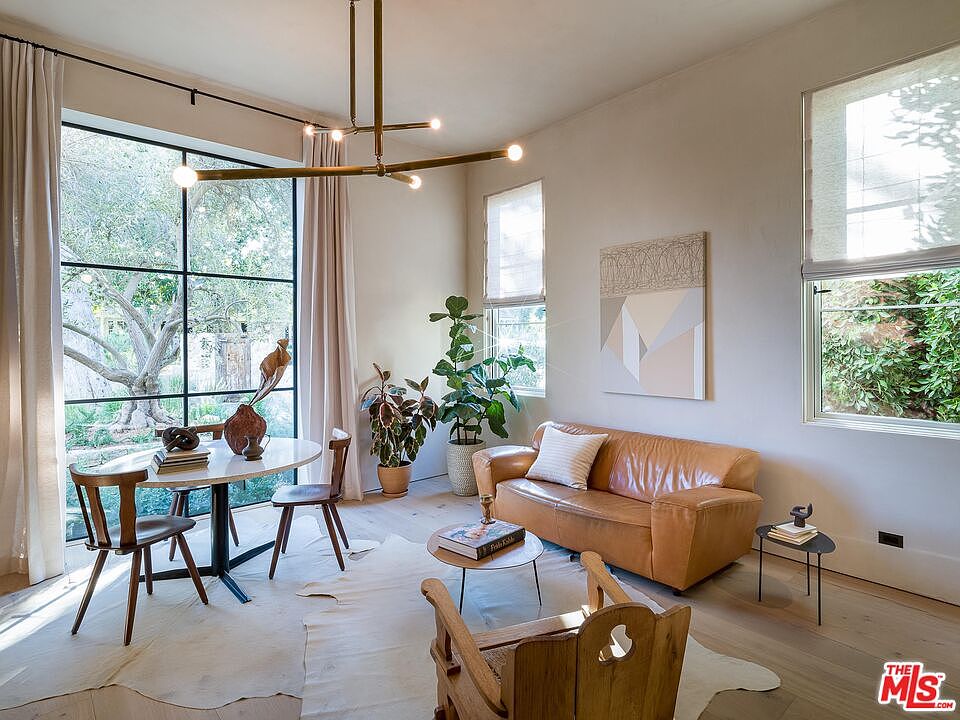 This interior shot showcases a stylish living room with a blend of modern and rustic elements. A leather sofa sits adjacent to a round coffee table and a wooden chair, while a dining area with a round table and chairs is positioned near a large window offering natural light and views of the outdoors. The room is decorated with plants and artwork, creating a warm and inviting atmosphere.
