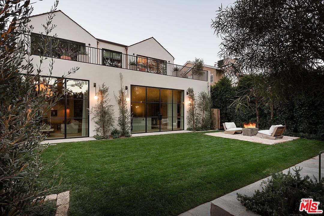 This image showcases the rear exterior of a modern home, emphasizing the well-manicured lawn and outdoor living space. Large glass doors provide seamless indoor-outdoor flow, while a fire pit area with seating offers a cozy gathering spot. The upper level features a balcony with black railings, adding architectural interest.