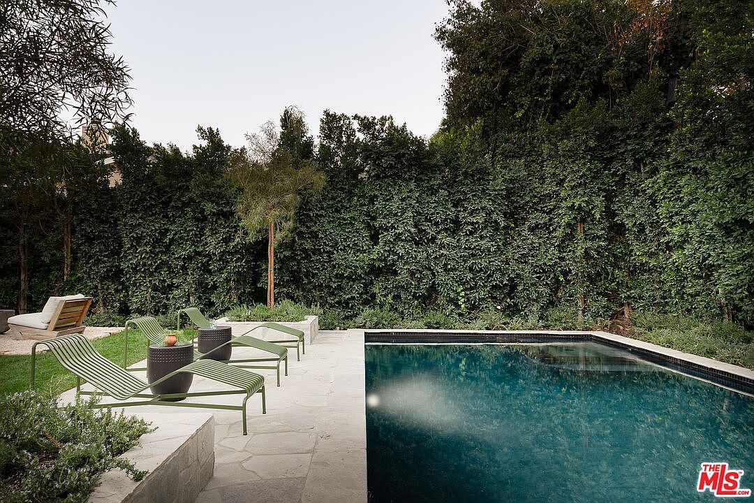 This image showcases a serene backyard pool area, featuring a rectangular pool with a tiled edge, surrounded by a stone patio. Several modern lounge chairs are arranged alongside the pool, complemented by small side tables. Lush greenery and tall hedges create a private and tranquil atmosphere, enhancing the property's appeal for relaxation and outdoor entertainment.