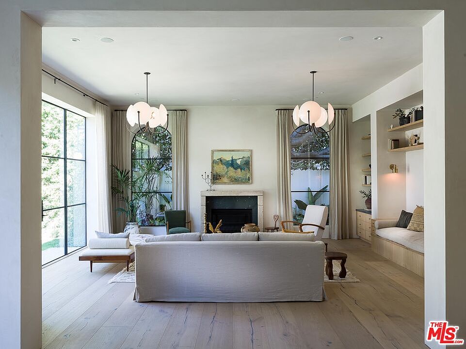 This is an interior shot of a living room featuring a large sofa, two chandeliers, and arched windows with curtains. The room has light hardwood floors and a fireplace, creating a bright and airy atmosphere. A built-in seating area with shelves is visible on the right side of the room.