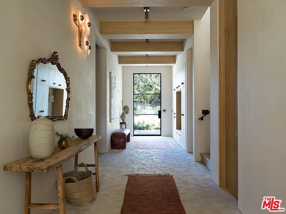 This interior shot showcases a hallway with a rustic-chic aesthetic. The hallway features exposed wooden beams, a textured wall finish, and a brick-patterned floor. A wooden console table with a decorative mirror and a woven basket adds character, while a black-framed glass door at the end of the hallway provides natural light and a view to the outside.