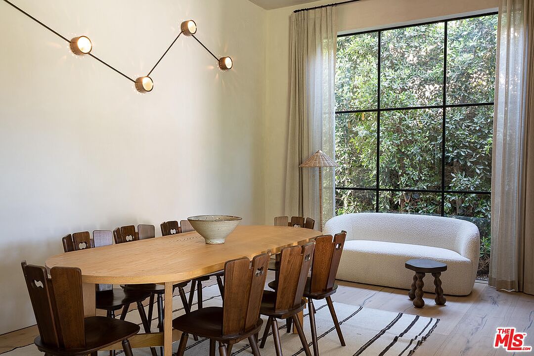 This dining room features a long, oval wooden table surrounded by wooden chairs with unique back designs. A modern, multi-bulb light fixture hangs above the table, and a large window with black frames offers a view of lush greenery outside. A cozy white sofa sits near the window, creating a comfortable and inviting atmosphere.