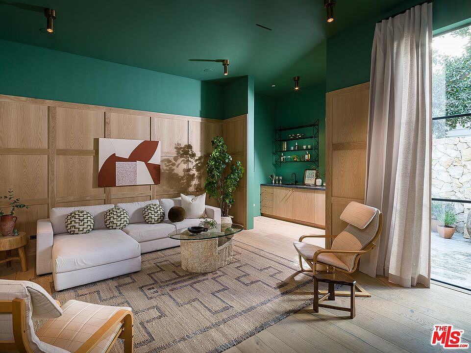 This interior shot showcases a stylish living room with a green ceiling and light wood paneling. A white sectional sofa is adorned with decorative pillows, complemented by a unique coffee table and a patterned rug. The room features a bar area with dark green walls and open shelving, adding a touch of sophistication.