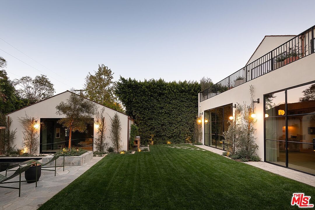 This image showcases a beautifully landscaped backyard featuring a lush green lawn, modern architectural elements, and mature trees. The yard is framed by a contemporary house with large windows and a separate structure, possibly a guesthouse or studio. The overall impression is one of tranquility and sophisticated outdoor living.