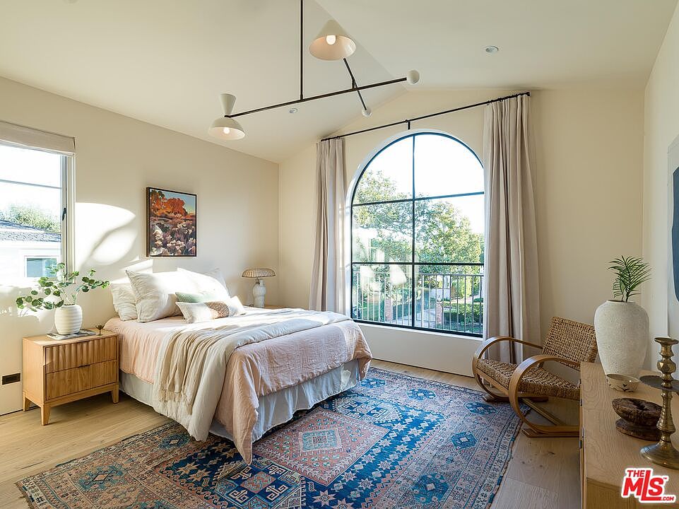 This is a bright and airy primary bedroom featuring a large arched window that provides ample natural light and a view of the outdoors. The room is decorated with a blend of modern and bohemian styles, including a patterned rug, a wooden rocking chair, and a unique overhead light fixture. The bed is neatly made with soft linens and a decorative throw, creating a cozy and inviting atmosphere.