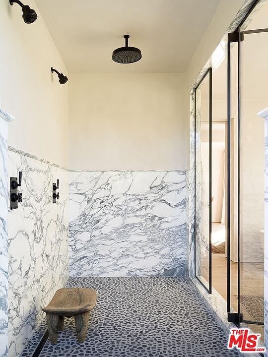 This is a luxurious primary bathroom featuring a walk-in shower with marble walls and a pebble stone floor. The shower is equipped with multiple shower heads, including a rainfall shower head. A rustic wooden stool adds a touch of warmth to the modern design, and the glass enclosure allows natural light to filter through.