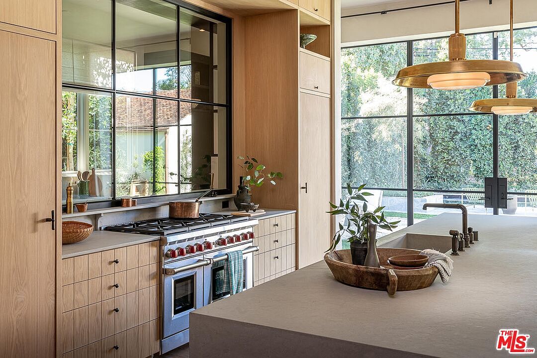 This is a well-lit kitchen featuring custom wood cabinetry and a large island with a concrete countertop. A professional-grade range is visible, and large windows offer views of the exterior greenery. The overall design is modern and sophisticated, with a focus on natural materials and clean lines.