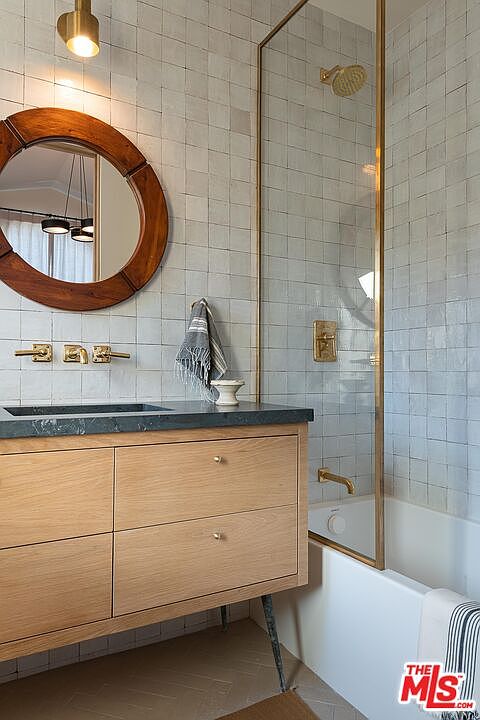 This is a well-lit primary bathroom featuring a wooden vanity with a dark countertop and a round wooden-framed mirror. The walls are tiled in a square pattern, and a glass-enclosed shower/tub is visible to the right. The overall style is modern and clean, with gold fixtures adding a touch of elegance.
