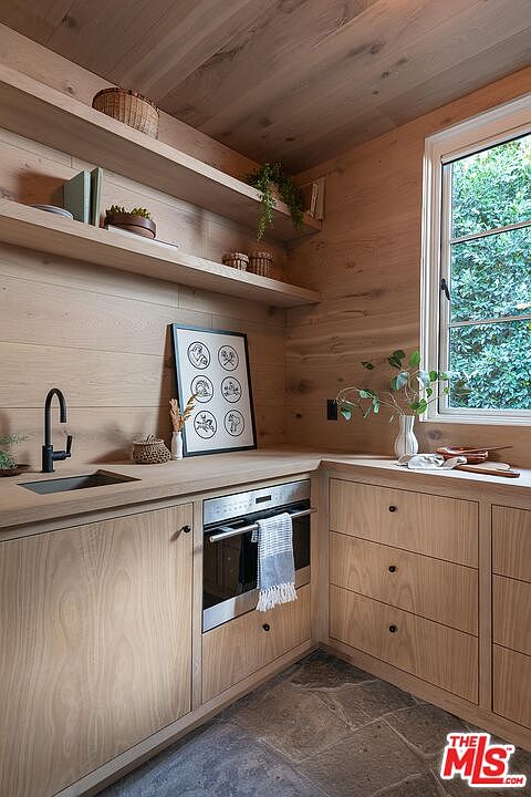 This interior shot showcases a modern kitchen with light wood cabinetry and countertops, complemented by a black faucet and stainless steel oven. Natural light streams in through a window, highlighting the clean lines and minimalist design. The space features open shelving and a framed art piece, adding a touch of personality.