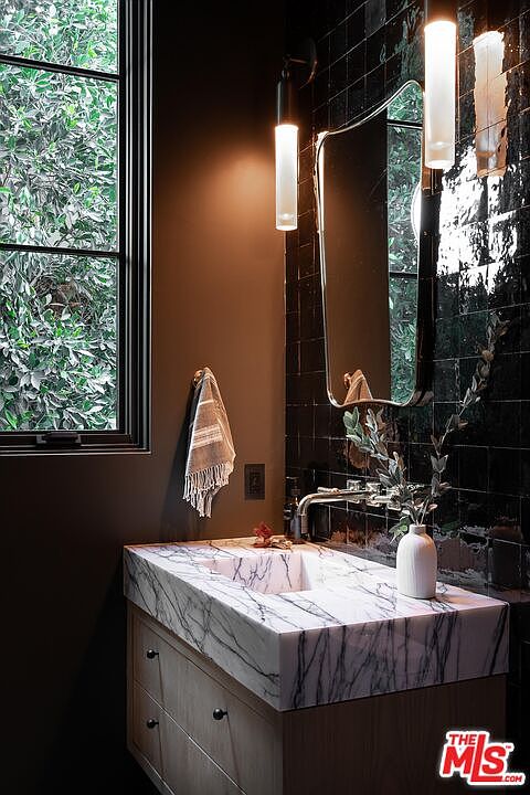 This bathroom features a modern aesthetic with dark tile walls and a striking marble countertop vanity. A sleek, rectangular mirror is illuminated by contemporary sconces, and a window provides natural light and a view of lush greenery. The overall impression is one of sophisticated elegance and tranquility.