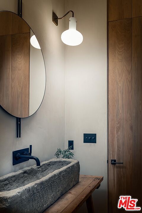 This image showcases a modern bathroom with a rustic touch. A stone trough sink sits atop a wooden table, complemented by a sleek black faucet and a round mirror. The lighting fixture adds a warm glow, highlighting the minimalist design and natural materials used throughout the space.