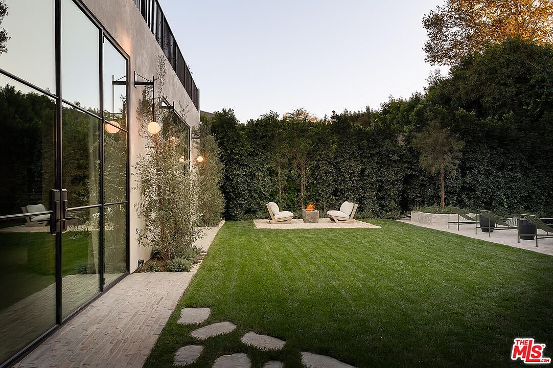 This exterior shot showcases a well-manicured backyard featuring a lush green lawn, a modern fire pit area with seating, and a sleek building facade with large windows. The landscape includes mature trees and hedges, providing privacy and a serene atmosphere. A stone pathway adds a touch of rustic charm, leading from the building to the lawn.