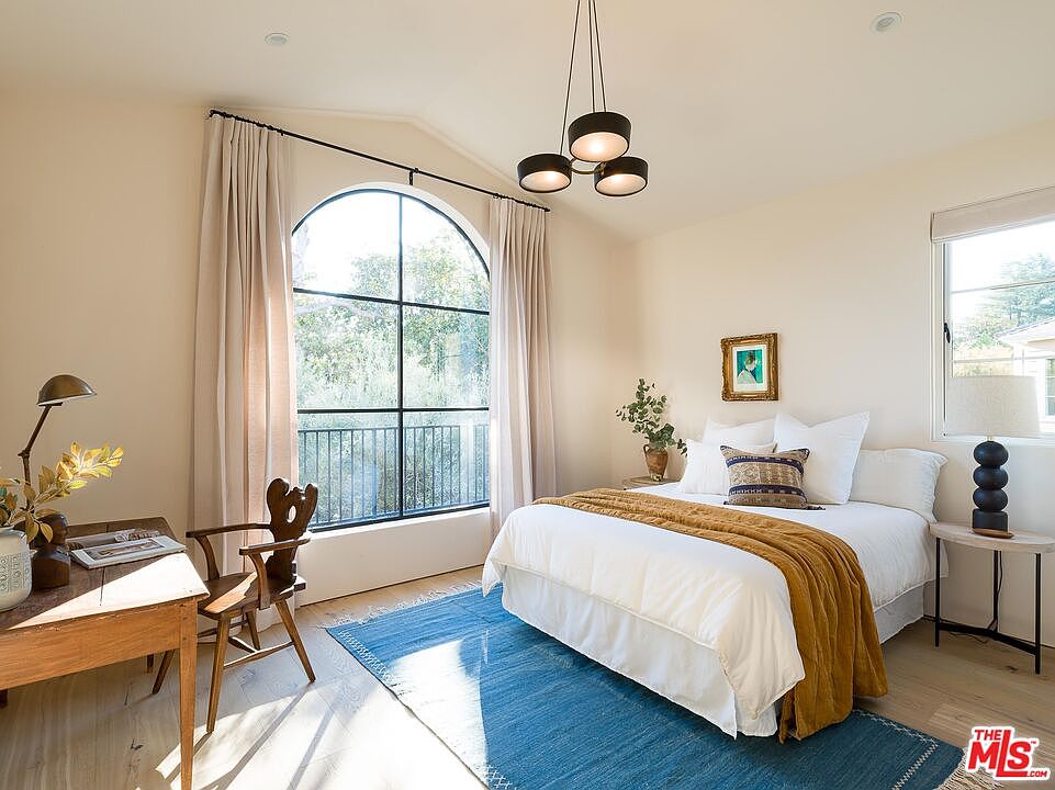 This is a bright and airy primary bedroom featuring a large arched window with natural light. The room is decorated in neutral tones with a blue rug, a wooden desk, and a bed with white linens and a mustard-colored throw. A modern chandelier hangs from the ceiling, adding a touch of contemporary style.
