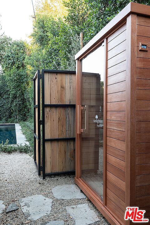 This image showcases a modern sauna in a backyard setting, featuring a wood-paneled exterior and a glass door with the "Sunlighten" logo. The sauna is situated on a gravel and stone pathway, with lush greenery and a glimpse of a pool in the background. The open wooden gate adds a touch of rustic charm to the scene.