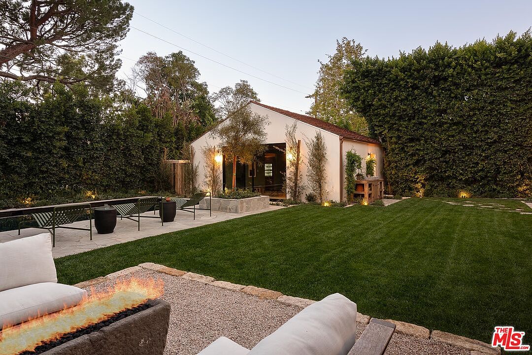 This image showcases a beautifully landscaped backyard featuring a lush green lawn, mature trees, and manicured hedges. A modern outdoor seating area with a fire pit adds a touch of luxury, while a charming outbuilding with large glass doors provides a focal point. The overall impression is one of tranquility and sophisticated outdoor living.