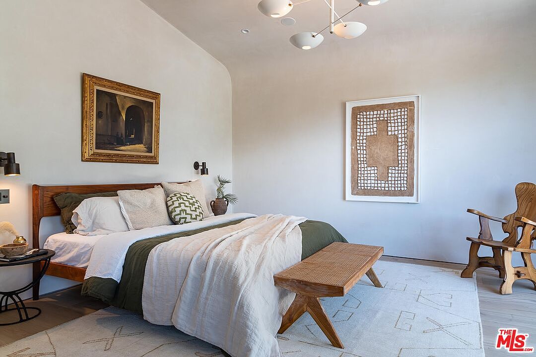 This is a serene primary bedroom featuring a wooden bed frame with neutral-toned bedding and decorative pillows. The room is adorned with artwork, including a framed painting and a modern piece, complemented by a wooden bench at the foot of the bed. The space is illuminated by a modern chandelier and wall sconces, creating a warm and inviting atmosphere.