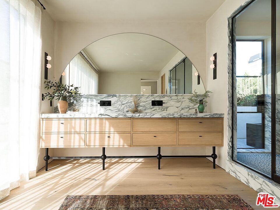 This primary bathroom features a double vanity with light wood drawers and a marble countertop, complemented by a large arched mirror. Black sconces flank the mirror, and natural light streams in through a nearby window, highlighting the light wood flooring. The overall design is modern and luxurious.