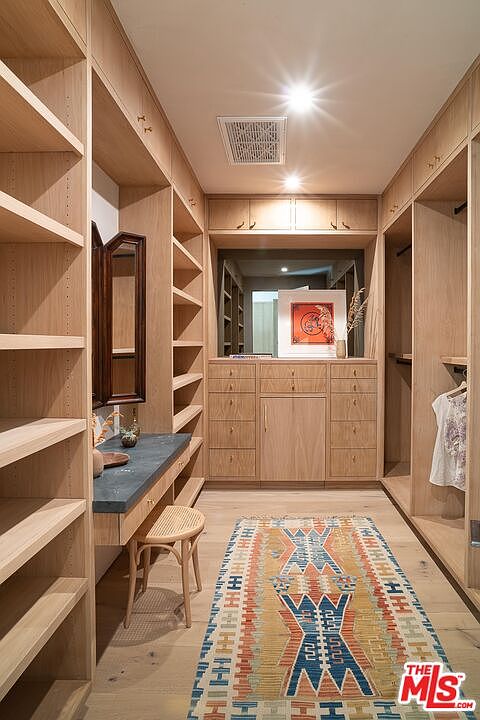 This is a well-organized walk-in closet featuring custom wood shelving and cabinetry. A vanity area with a dark countertop and a woven stool is present, and a colorful rug adds a touch of style to the hardwood floor. The closet is brightly lit, creating an inviting and functional space.