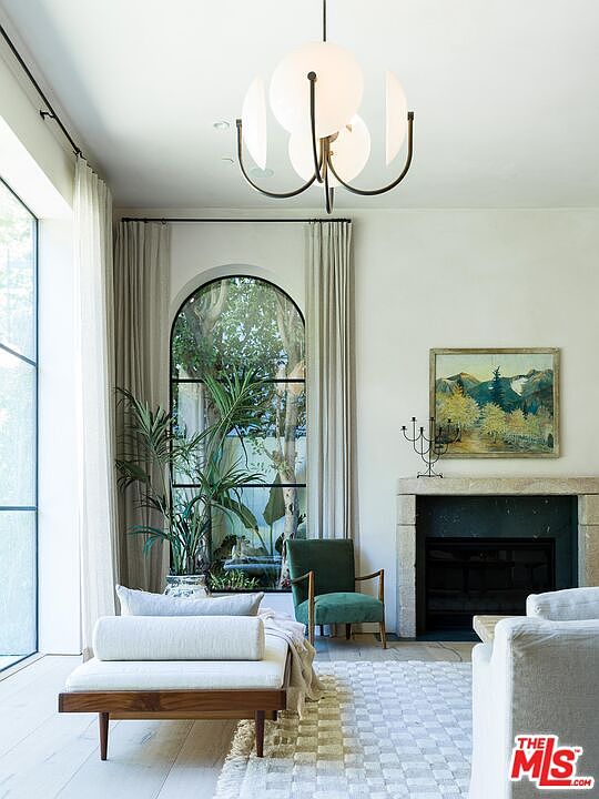 This is an interior shot of a living room featuring a modern aesthetic. The room is well-lit with natural light coming through a large arched window. Key elements include a daybed, a green accent chair, a fireplace with a stone surround, and a unique globe chandelier, creating a sophisticated and inviting atmosphere.
