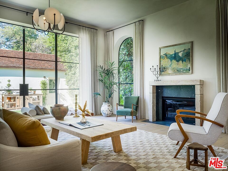 This is an interior shot of a living room featuring a large window with black frames, allowing natural light to flood the space. The room is furnished with a light-colored sofa, a wooden coffee table, and a stone fireplace with a painting above it. The overall style is modern and inviting, with a focus on natural materials and a neutral color palette.