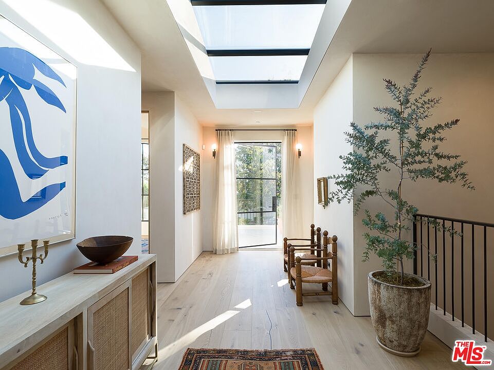 This interior shot showcases a bright hallway with a skylight, light wood flooring, and neutral-toned walls. The hallway features artwork, decorative furniture, and a potted tree near the staircase, creating a sophisticated and inviting atmosphere. A doorway at the end of the hall leads to an outdoor area, enhancing the sense of space and light.