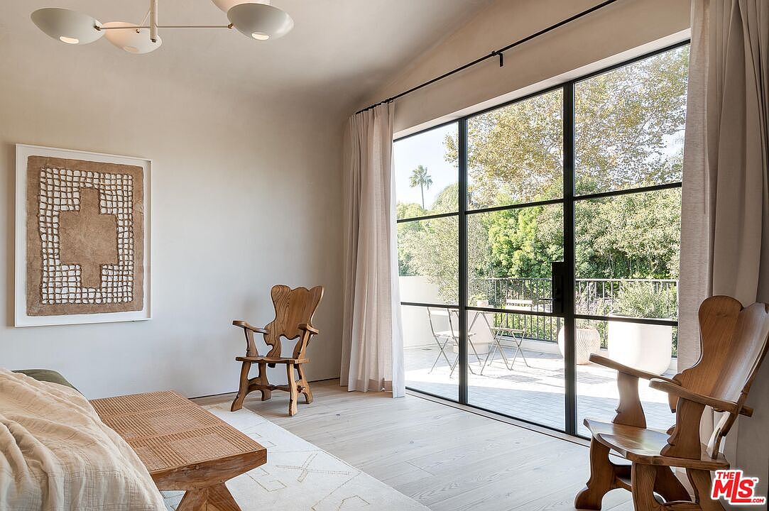 This interior shot showcases a bright and airy living room with a modern aesthetic. The room features light wood flooring, a large window with black frames offering an outdoor view, and neutral-toned walls. The decor includes wooden chairs, a woven bench, and an abstract art piece, creating a serene and stylish atmosphere.