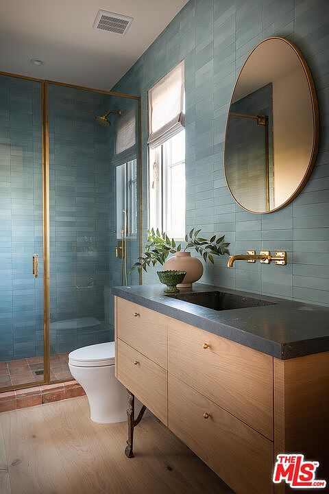 This is a stylish bathroom featuring blue-green tiled walls and a modern vanity with a dark countertop and light wood cabinetry. A large, round mirror hangs above the sink, and a glass-enclosed shower is visible to the left. The overall aesthetic is clean, contemporary, and luxurious.
