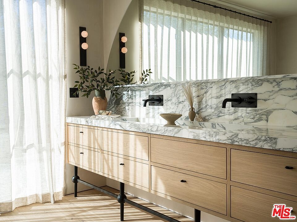 This is a bright and modern primary bathroom featuring a double vanity with a marble countertop and light wood cabinetry. Black fixtures, including wall-mounted faucets and sconces, provide a striking contrast. The room is illuminated by natural light filtering through sheer curtains, creating a serene and luxurious atmosphere.