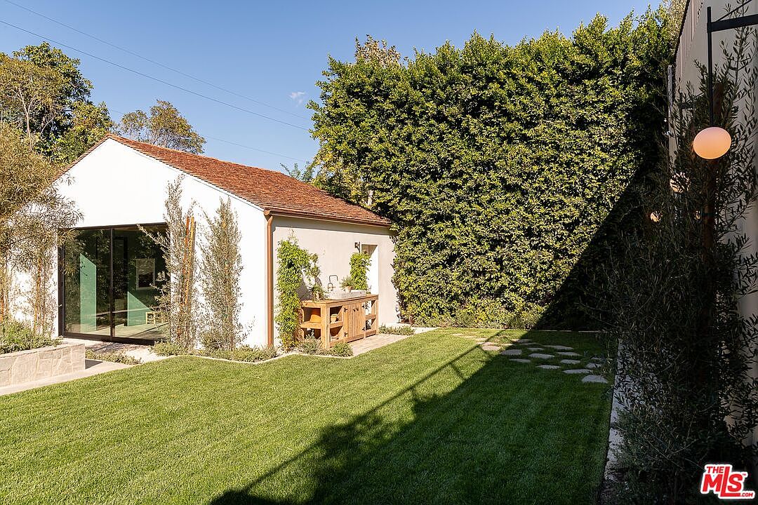 This image showcases a well-maintained backyard featuring a lush green lawn and a modern, minimalist building with large glass windows. A tall, dense hedge provides privacy, while a wooden outdoor kitchen area adds functionality. The yard is neatly landscaped with stepping stones and various plants, creating a serene and inviting outdoor space.