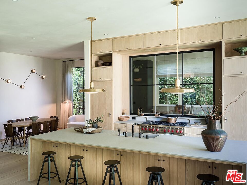 This is a bright and modern kitchen featuring light wood cabinetry and a large island with seating. The kitchen is well-lit by pendant lights and a large window that provides natural light and a view of the outdoors. The space also includes a dining area, creating an open and inviting atmosphere.