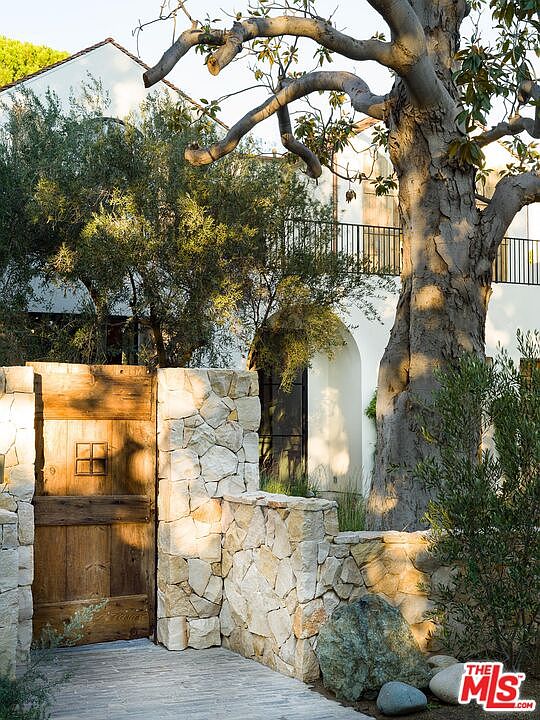 This image showcases a charming entryway featuring a rustic wooden gate set within stone walls. A large, mature tree provides shade and adds to the natural ambiance. The pathway leads towards a white building with an arched doorway and a balcony with black railings, creating an inviting and picturesque scene.