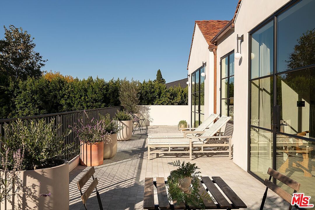 This image showcases a stylish patio or deck area, featuring modern outdoor furniture including lounge chairs and a wooden table with chairs. The space is adorned with potted plants, adding a touch of greenery and sophistication. Large windows with black frames provide a seamless transition to the interior, enhancing the indoor-outdoor living experience.