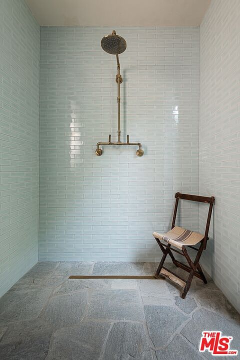 This is a bathroom featuring light blue subway tile walls and a stone tile floor. A vintage-style shower fixture is mounted on the wall, and a wooden folding chair sits in the corner. The perspective is from the doorway, showcasing the entire shower area.