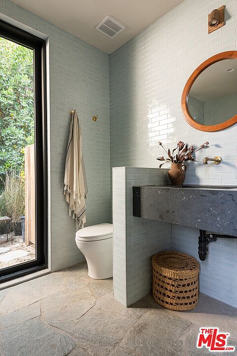This is a well-lit guest bathroom featuring light blue subway tile walls and stone flooring. A modern floating vanity with a dark countertop is complemented by a round wooden-framed mirror. The room includes a toilet and a window offering a view of the outdoors, creating a serene and stylish space.