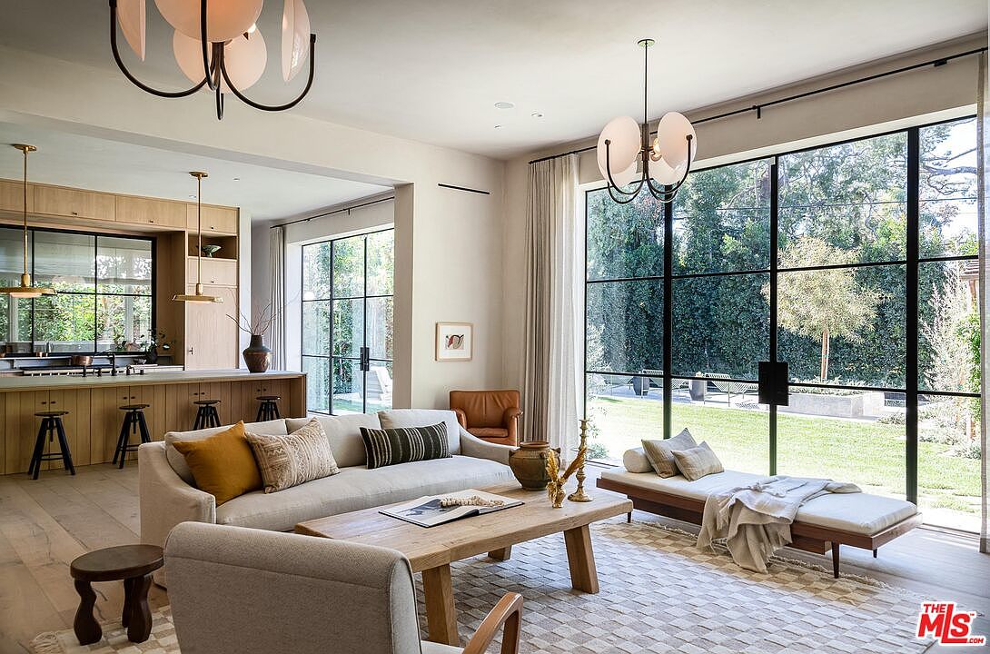 This is a bright and airy living room featuring a neutral color palette, large windows with black frames, and hardwood floors. The room is furnished with a comfortable sofa, a wooden coffee table, and a daybed near the window, creating a cozy and inviting atmosphere. The open floor plan connects seamlessly to the kitchen area, enhancing the sense of spaciousness.